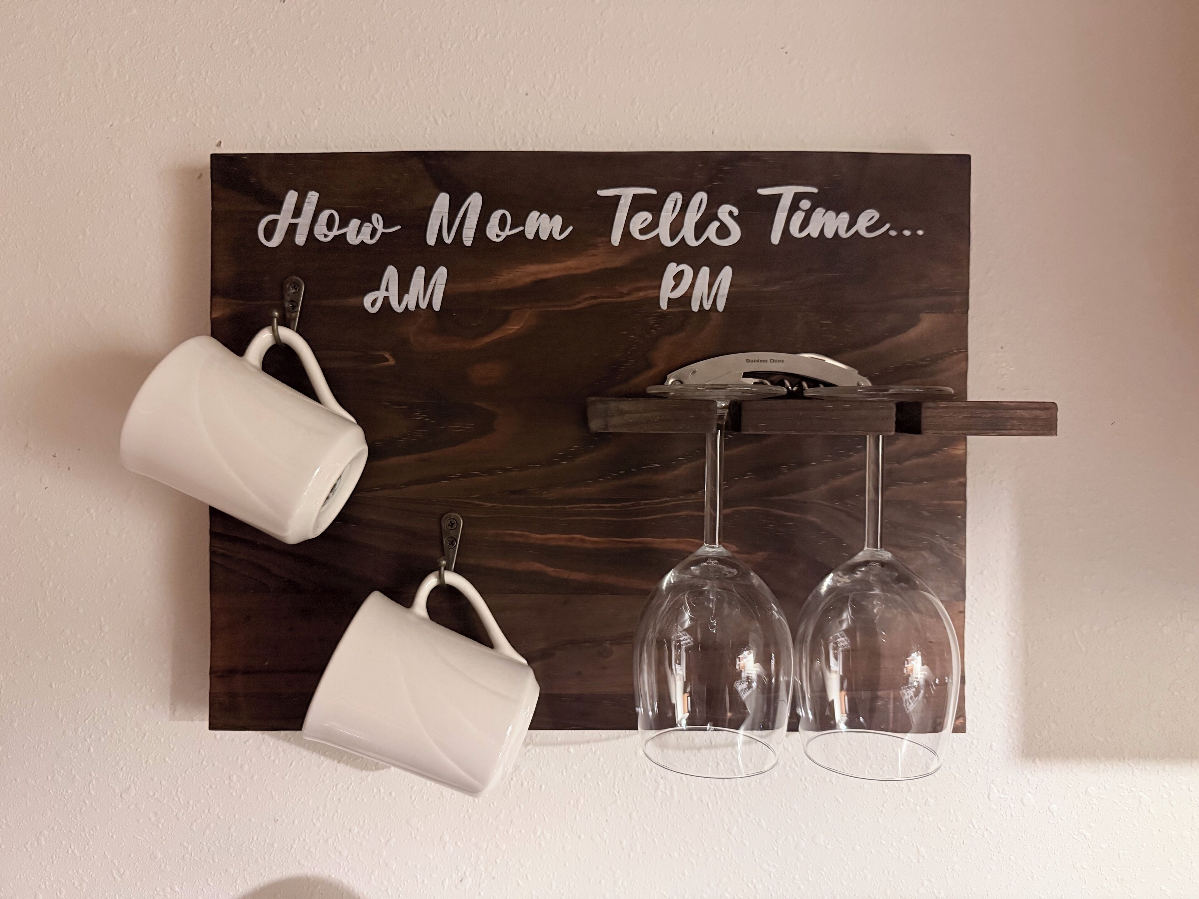 Une horloge murale en bois sur laquelle est inscrit « Comment maman apprend à lire l’heure… matin et soir », fixée au mur, avec deux tasses blanches et deux verres à vin suspendus en dessous.