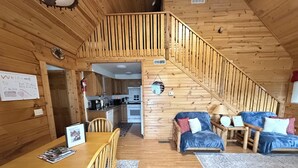 A cabin interior with a wooden staircase, a dining area with a table and chairs, a kitchen, and a living area with a sofa and a lamp.