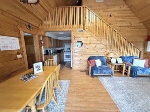 Ein Blockhaus-Interieur mit einer Holztreppe, einem Essbereich mit Tisch und Stühlen, einer Küche und einem Wohnbereich mit Sofa und Lampe.