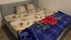 Een kleine kamer met een enkel bed, een grijs hoofdeinde, een bloemenbedekking en een blauwe deken met een