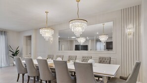 Une salle à manger avec une grande table rectangulaire, six chaises rembourrées et trois lustres en cristal.