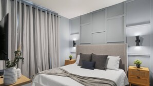 Une chambre moderne avec un grand lit, des rideaux gris, une télévision, des tables de chevet avec des lampes et une étagère fixée au mur.