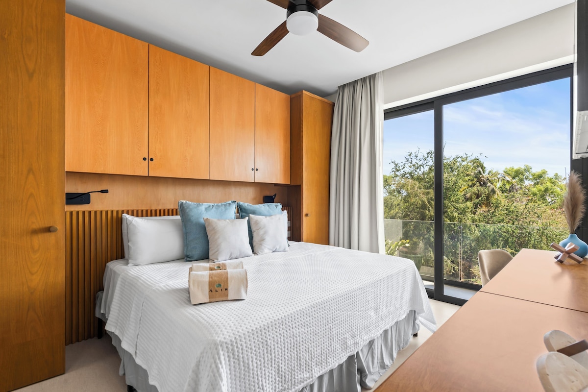 Ein Schlafzimmer mit einem großen Bett, Holzkombinationen, einem Deckenventilator, einem Tisch und Blick ins Grüne.