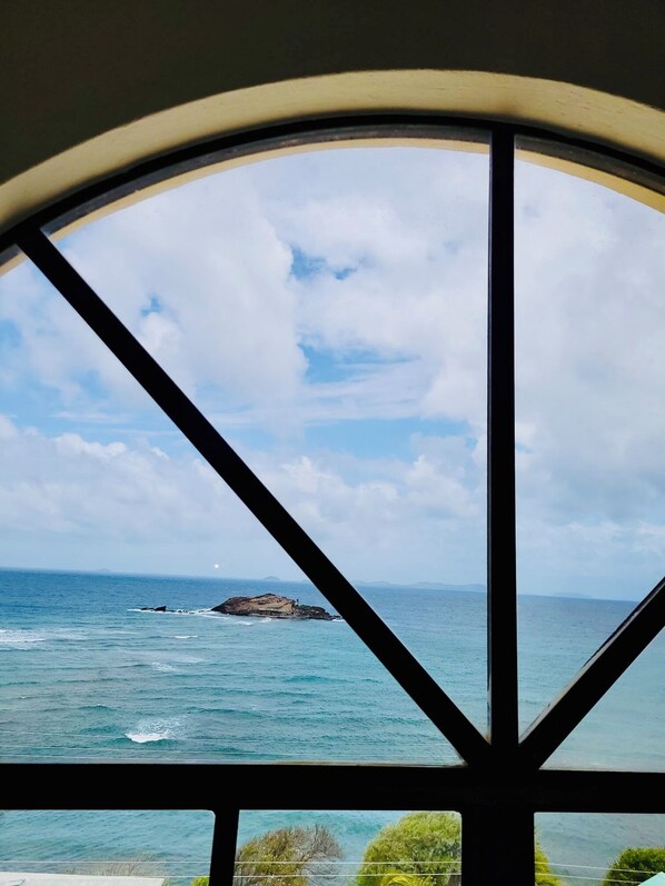 Une vue à travers une fenêtre à cadre incurvé, donnant sur une zone côtière avec un promontoire rocheux et une mer calme.