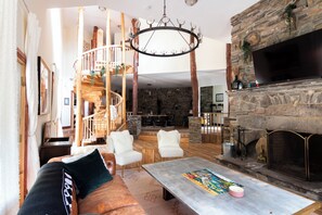 A spacious living room with a stone fireplace, a large TV, and a wooden staircase leading to an upper level.