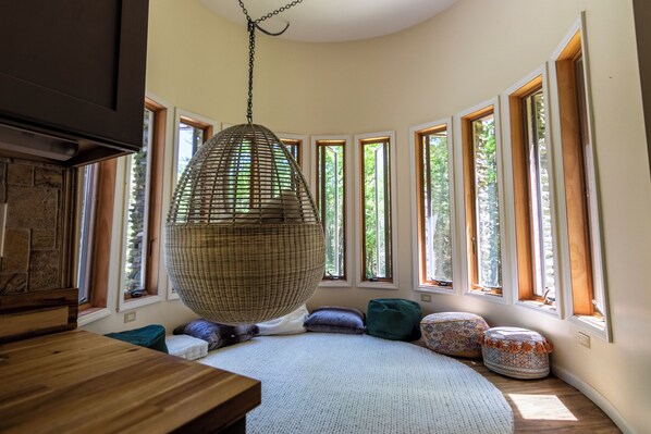 A cozy room with a large woven hanging chair, a wooden table, and multiple windows offering views of greenery.