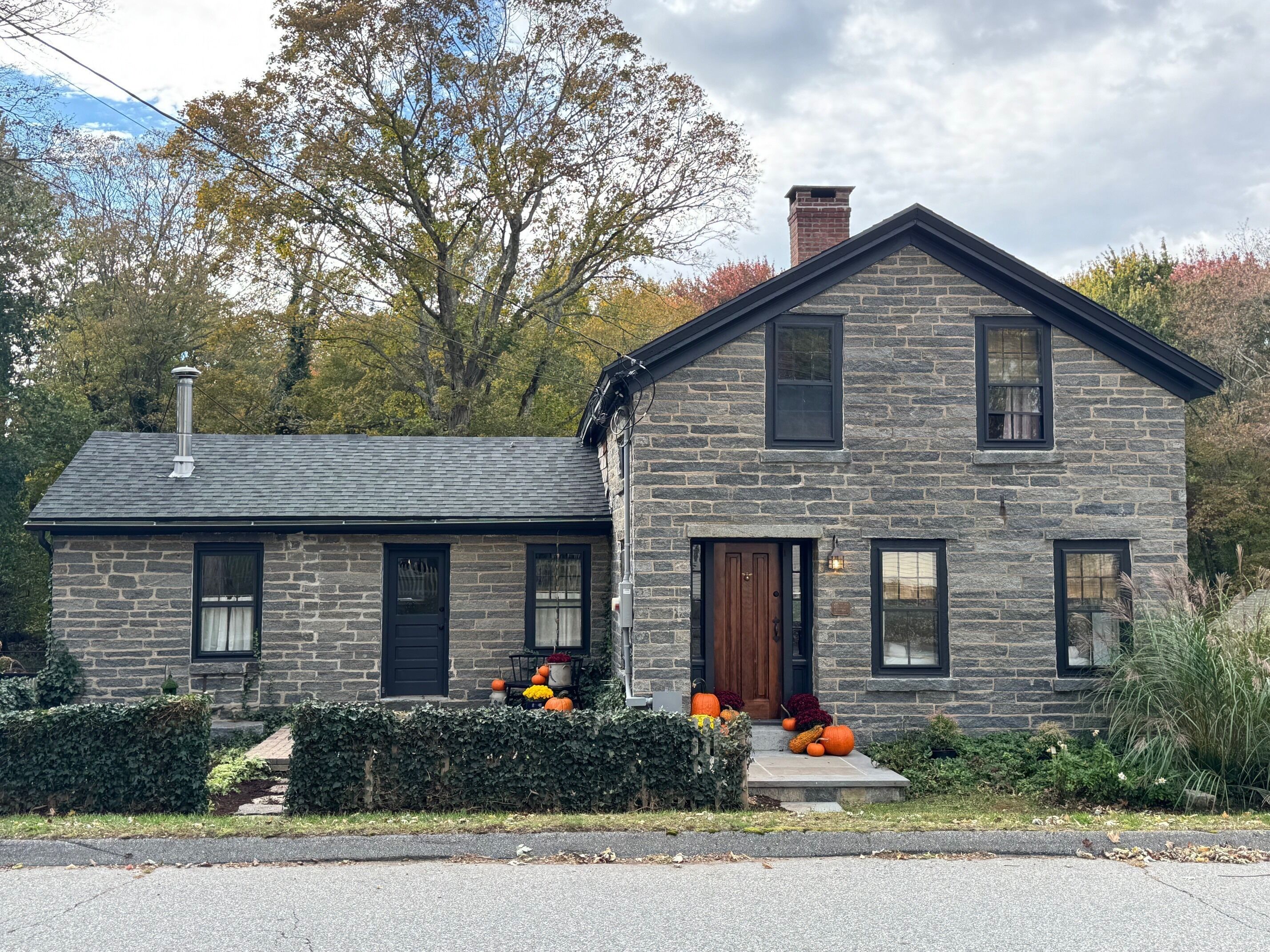 Une maison en pierre avec un toit sombre, une cheminée et une porte d’entrée. On peut voir des citrouilles et d’autres décorations d’automne sur le porche.