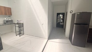 A kitchen with white walls, a refrigerator, a microwave, and a small table with a box on it. - Departamento Residencial San Juan Bosco  (Guadalajara)