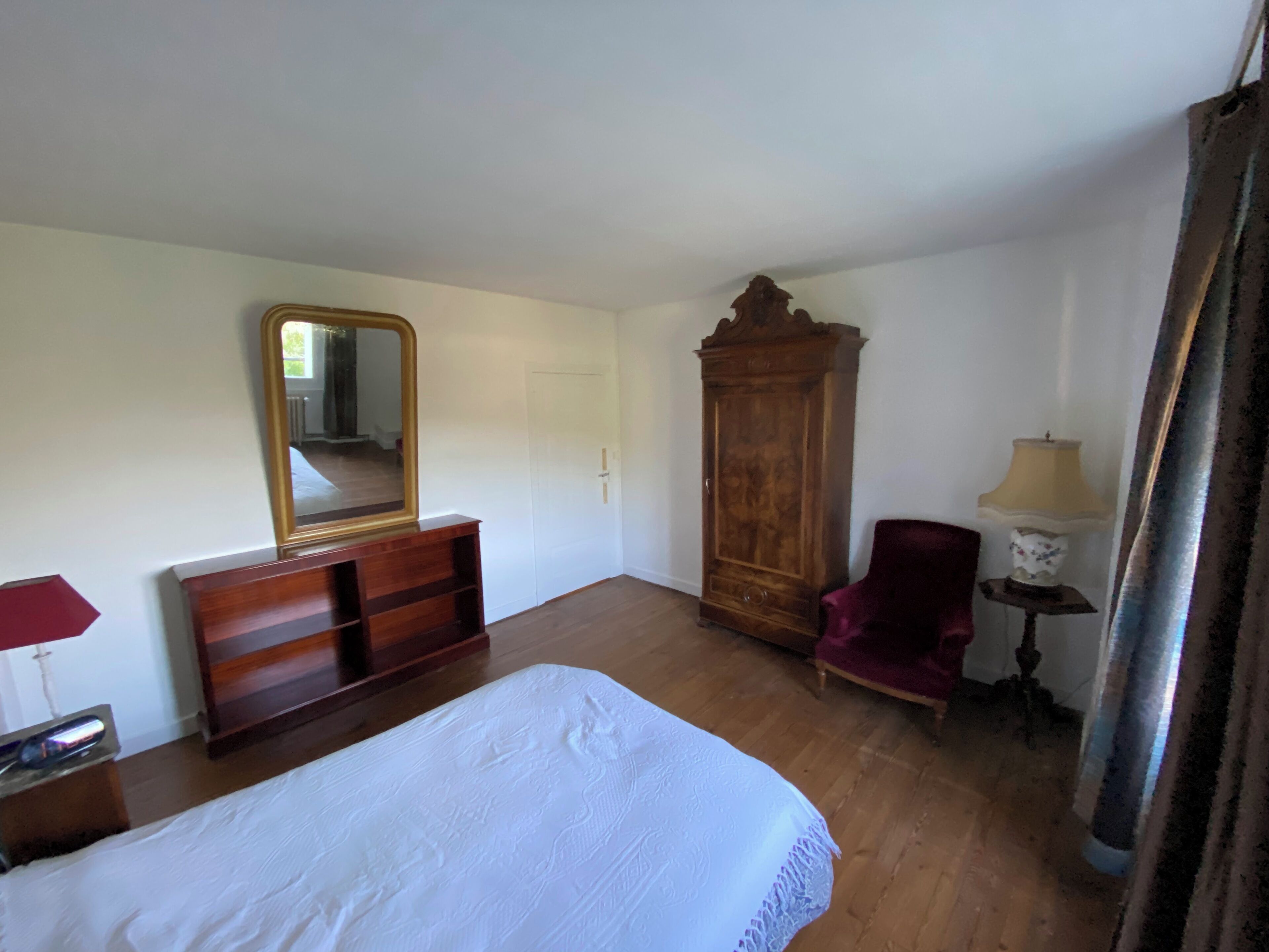 A bedroom with a bed, dresser, mirror, wardrobe, chair, lamp, and curtains.