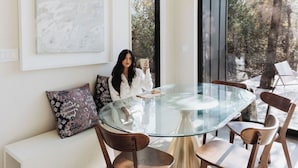 A modern dining area with a glass table and wooden chairs, a white sofa with patterned cushions, and a large abstract painting on the wall.