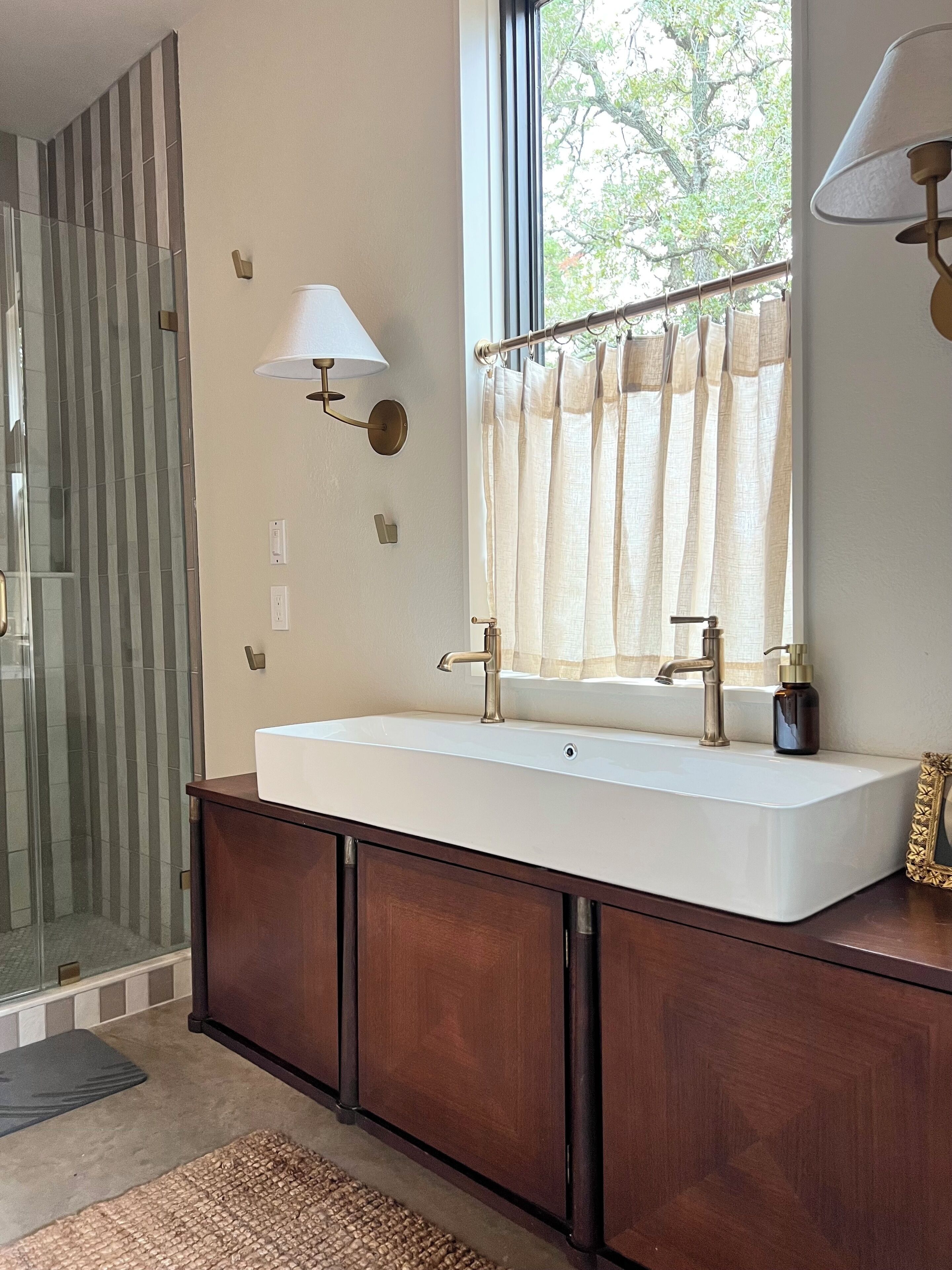 A bathroom with a large white sink, a mirror, a window with a curtain, and a wall-mounted light fixture.