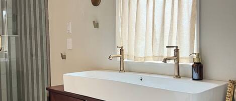 A bathroom with a large white sink, a mirror, a window with a curtain, and a wall-mounted light fixture.