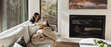 A modern living room with a large window, a fireplace, and a painting of a mountain landscape.