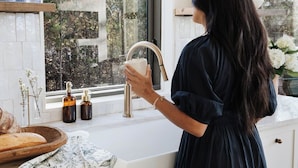 A person in a dark dress at a kitchen counter with a window, a sink, and a wooden cutting board with bread.