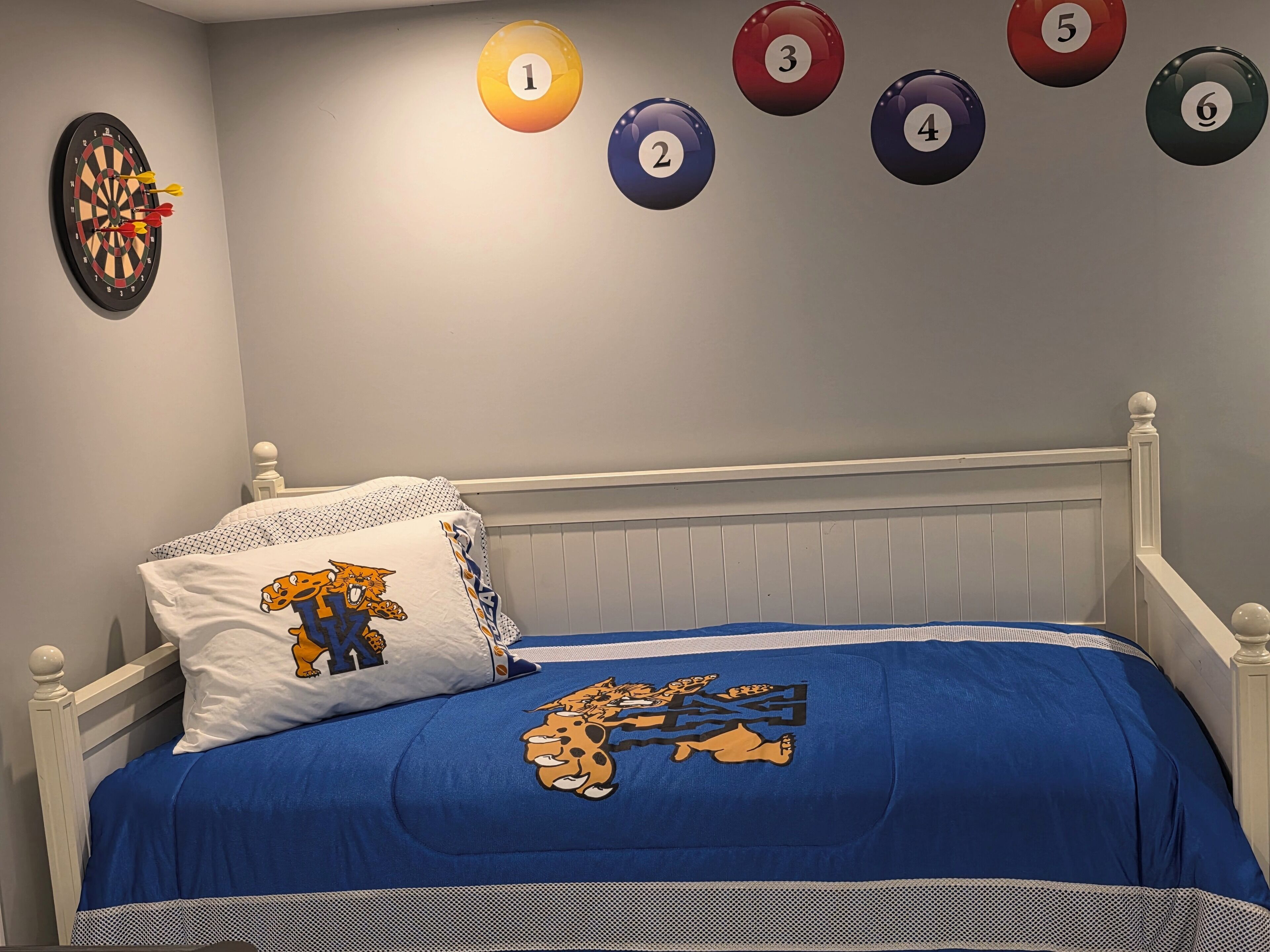 Un dormitorio con cama, un almohadón con personaje de caricatura, una colcha azul, un dardo y una mesa de billar con bolas numeradas.