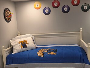 Un dormitorio con cama, un almohadón con personaje de caricatura, una colcha azul, un dardo y una mesa de billar con bolas numeradas.