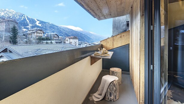 Ein Balkon mit Blick auf schneebedeckte Berge, einem hölzernen Hocker und einem Buch auf einer Ablagefläche.