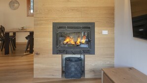 A modern living room with a wood-paneled wall, a built-in fireplace with visible flames, a dining table with chairs, and a TV mounted on the wall. - AlpinWood - Two Bedroom Apartment, Sleeps 5 (Temù)