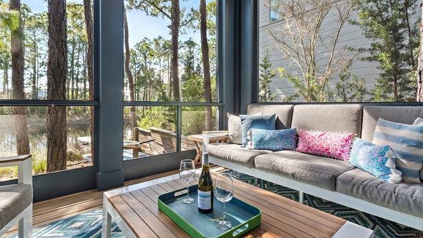 Ein gemütliches Zimmer mit großem Fenster, einer Couch mit bunten Kissen, einem Holztisch mit Weinflasche und Gläsern sowie Blick auf Bäume und ein Gewässer.