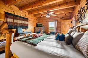 A log cabin interior with a bed, a television, a fireplace, and a ceiling fan. - Mistwood Manor | Mtn Views • Hot Tub • Game Room (Sevierville)