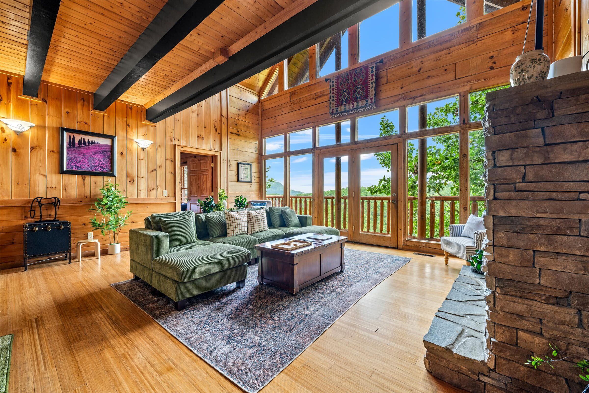 A spacious living room with a large window, wooden walls, a green sofa set, a coffee table, and a stone fireplace.