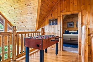 A wooden-floored room with a foosball table, a bed with a blue and white headboard, and a wooden railing. - Mistwood Manor | Mtn Views • Hot Tub • Game Room (Sevierville)