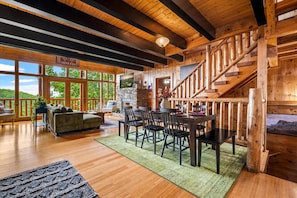 A spacious living area with a fireplace, wooden staircase, and large windows offering a view of the outdoors. - Mistwood Manor | Mtn Views • Hot Tub • Game Room (Sevierville)
