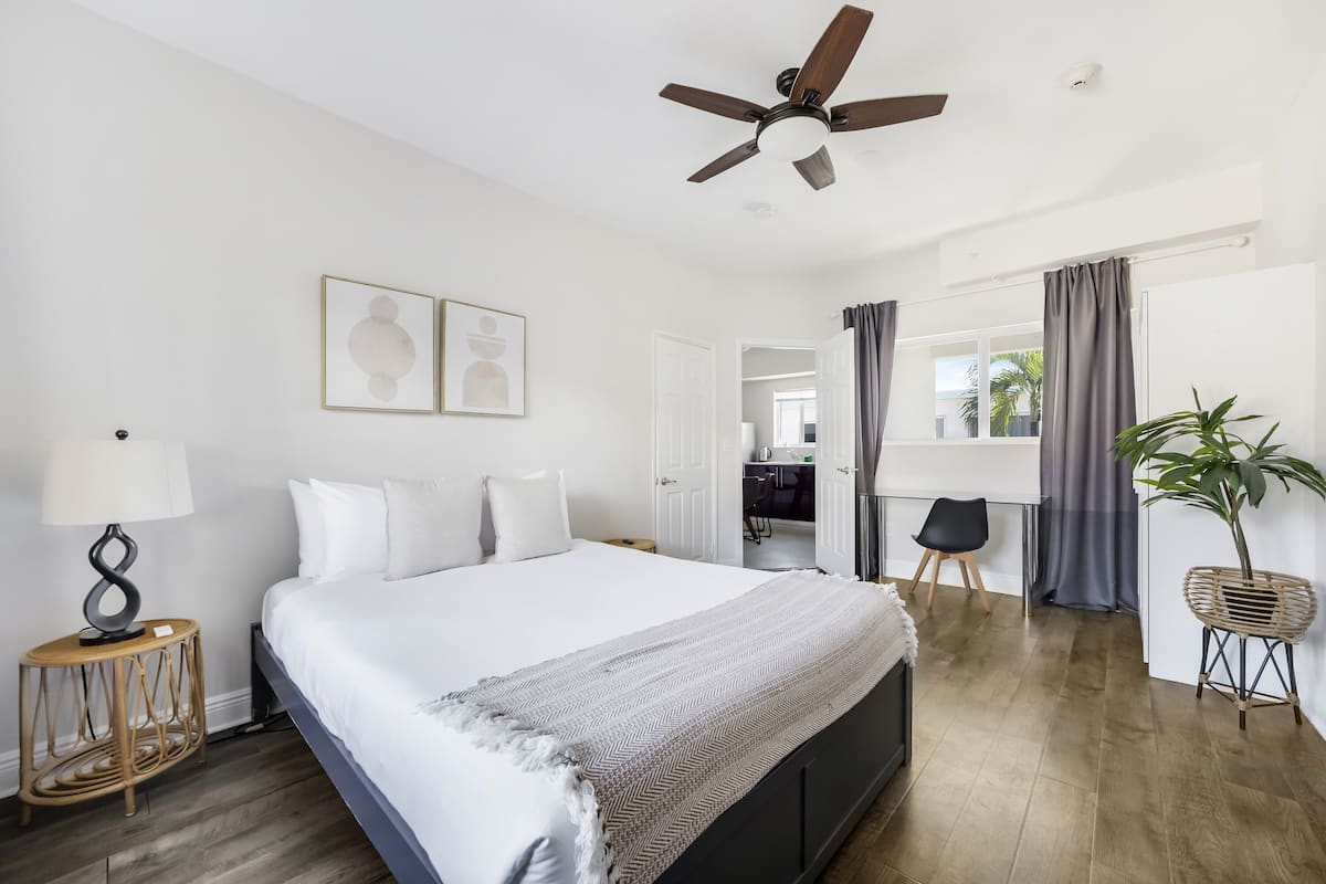 A modern hotel room with a large bed, a ceiling fan, a bedside table with a lamp, and a view of the outside through a window with curtains.