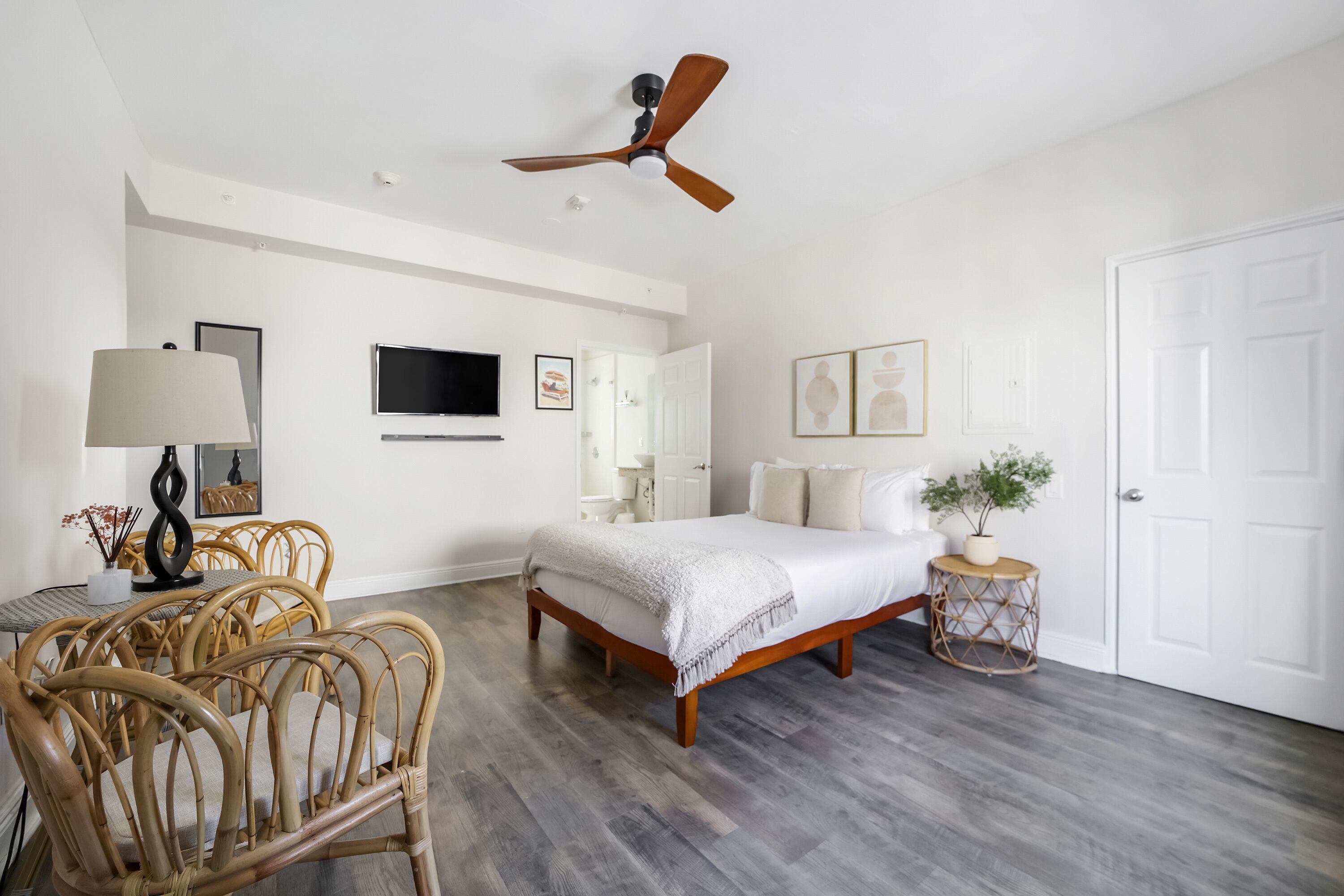 A bedroom with a bed, a ceiling fan, a TV, a dining table with chairs, and a small round table with a plant.