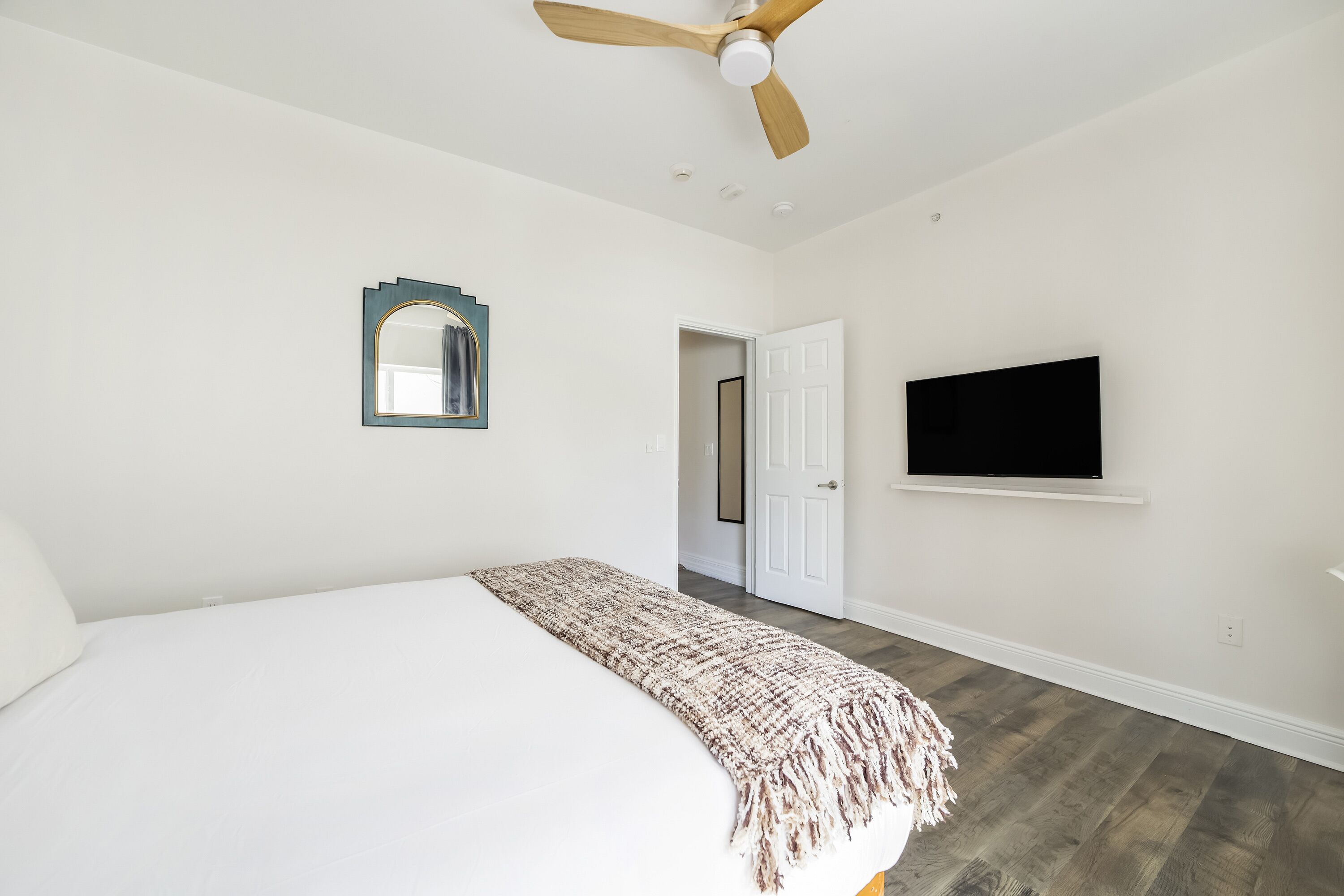 A bedroom with a bed, a ceiling fan, a television, and a door leading to another room.