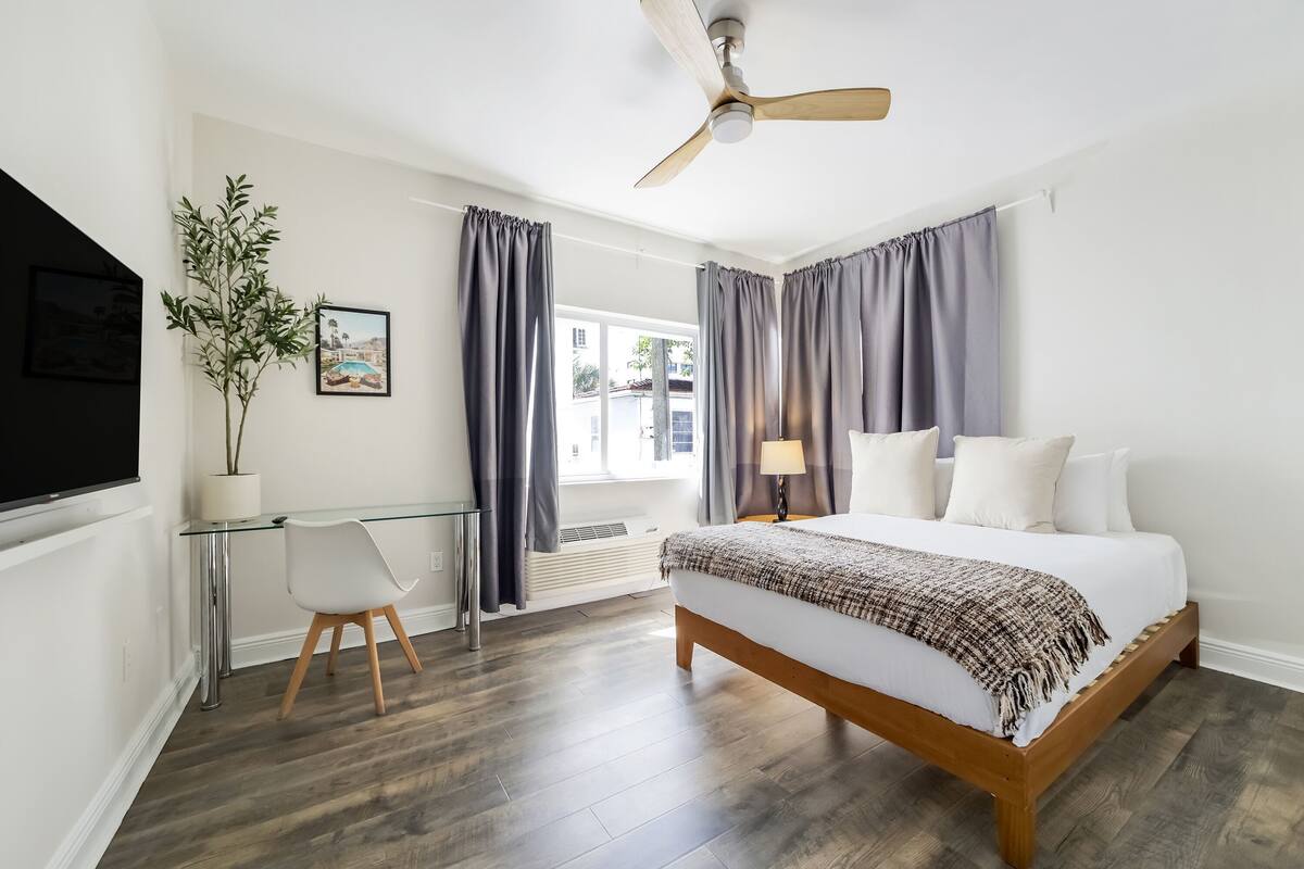 A bedroom with a bed, a desk, a chair, a ceiling fan, and a window with curtains.