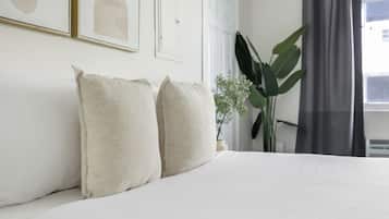 Una cama bien hecha con ropa blanca, una planta verde grande y cuadros en la pared.