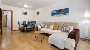 A living room with a sofa, a dining table with chairs, a lamp, and artwork on the walls. - ‘Ria Formosa Pool Heaven’ Apartment with Shared Pool, Wi-Fi, and Air Conditioning (Olhão)