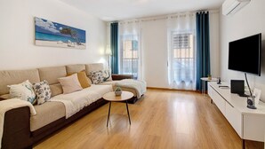 A living room with a sofa, a coffee table, a TV, and a painting of a beach scene. - ‘Ria Formosa Pool Heaven’ Apartment with Shared Pool, Wi-Fi, and Air Conditioning (Olhão)