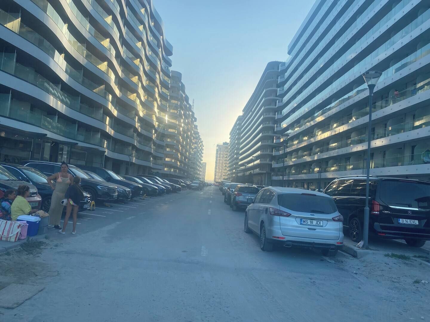 En parkeringsplads med flere biler og nogle mennesker, der står i nærheden af indgangen.