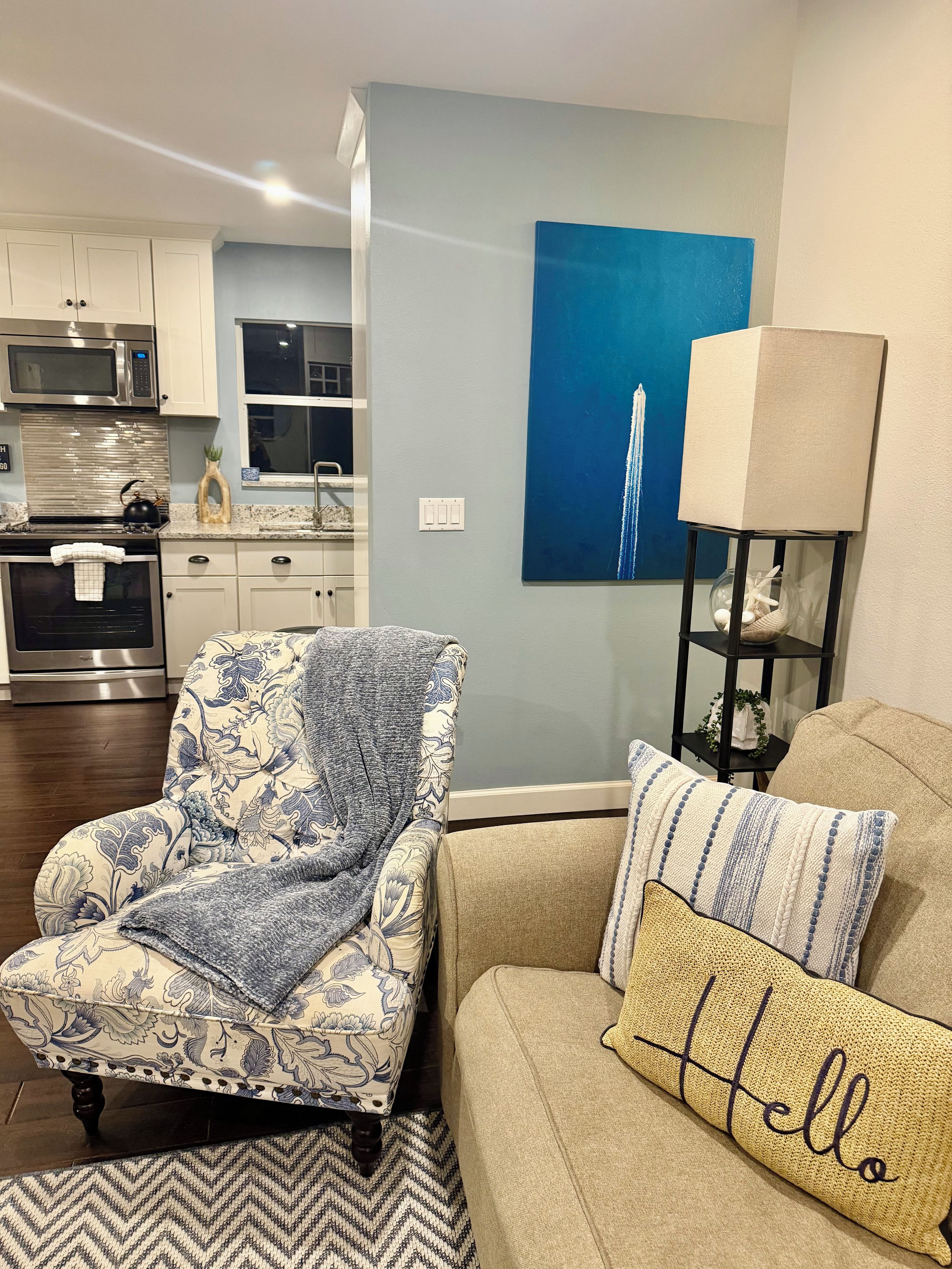 Un salón con un sillón estampado, un sofá con un cojín que dice 'Hello', una lámpara y un cuadro de una estructura azul.