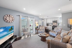 A modern living room with a flat-screen TV, a large wall clock, and a dining area with a kitchen in the background. - Cozy 2-bedroom condo with dock! Best location in Anna Maria. Beach & sunsets  (Anna Maria)