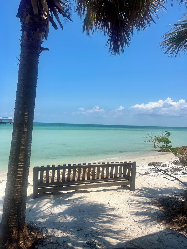 A bench on a sandy beach with clear turquoise water and a palm tree in the foreground.