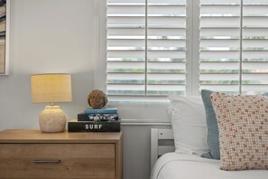 A neatly arranged bedroom with a bedside table, a lamp, a stack of books, and a window with blinds.