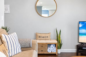 A living room with a sofa, a wooden side table with a beach-themed pillow, a round mirror, a potted plant, and a flat-screen TV. - 2BR Waterfront with Dock. Best area in Anna Maria. Beach, Sunsets on Pine Ave. (Anna Maria)