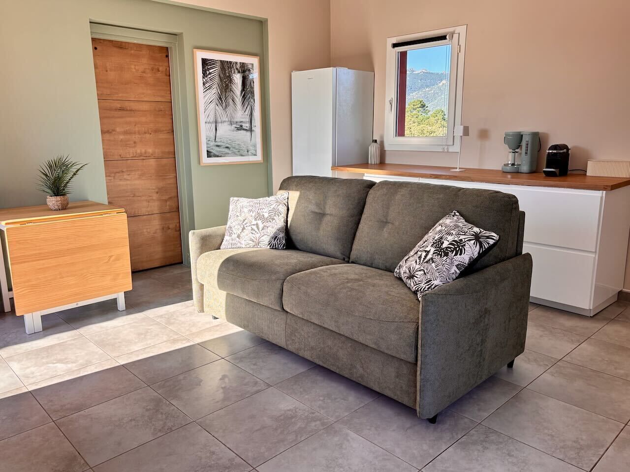 A modern living room with a sofa, a wooden side table, a white refrigerator, and a window with a view of greenery.