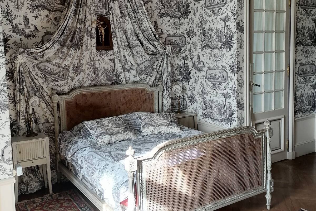 Ein Schlafzimmer mit Baldachinbett, gemustertem Wandpapier, Holzboden und einer Tür mit Glasfeldern.