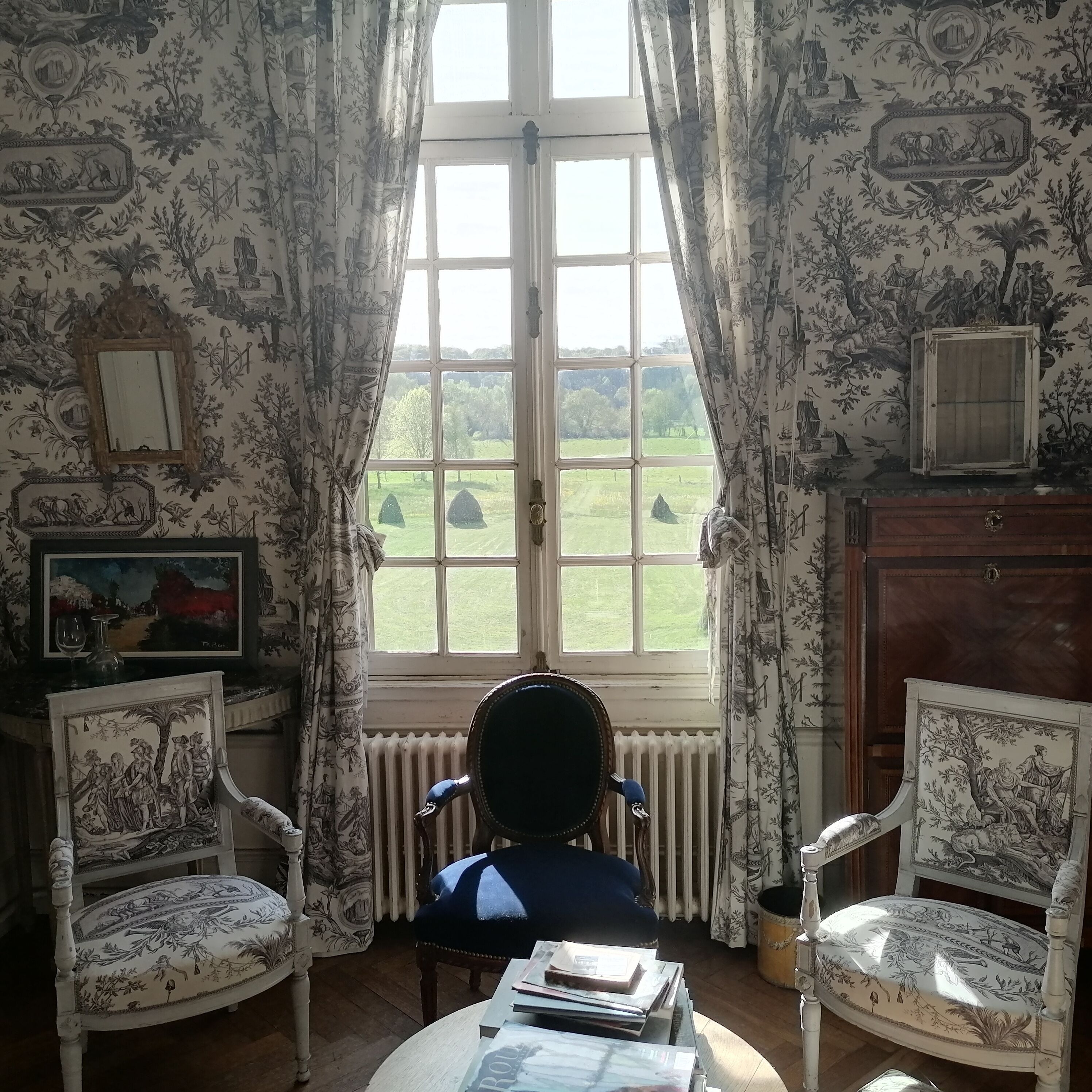A room with a large window, two chairs, a table with books, and a radiator.