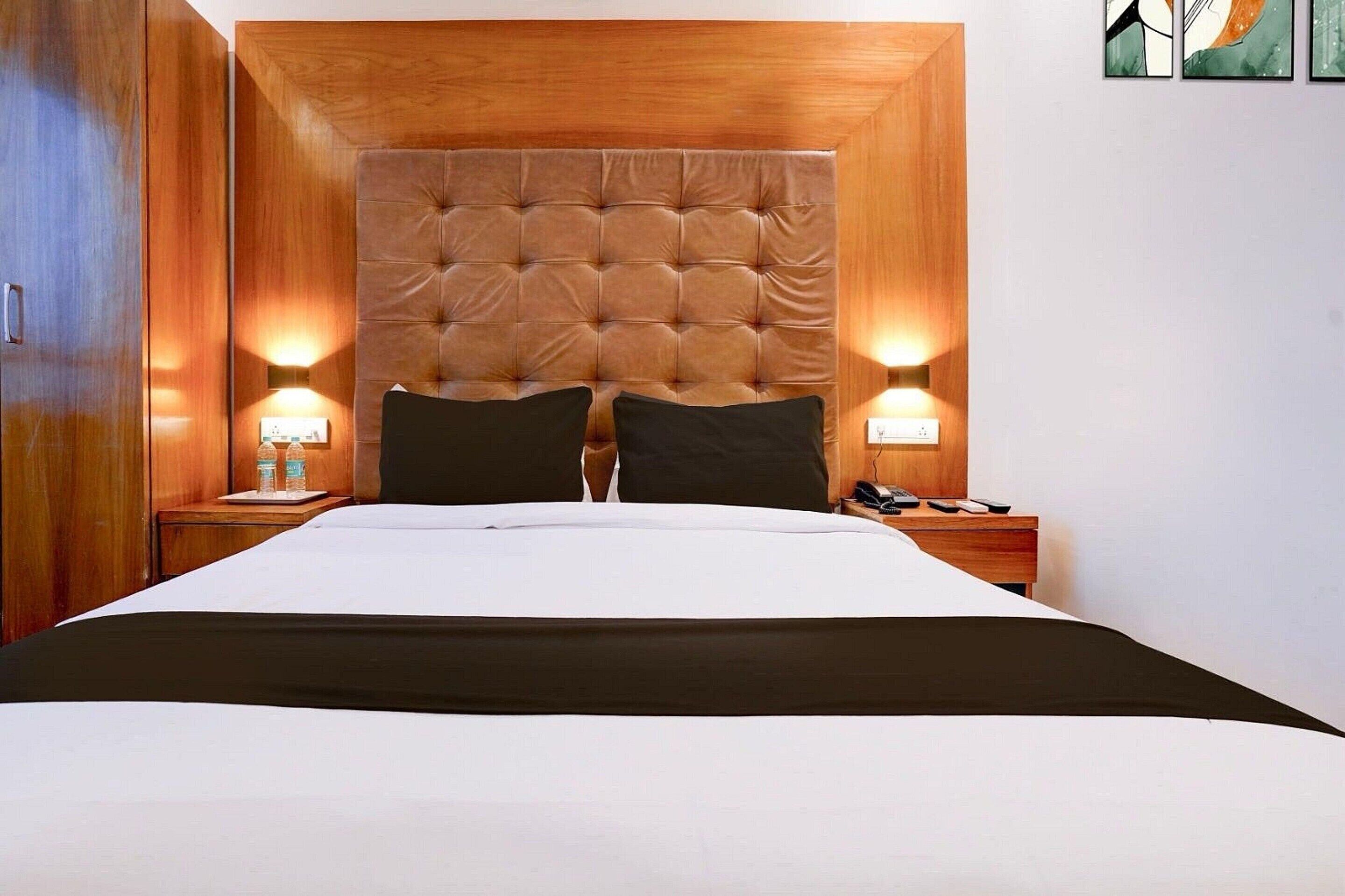 Una habitación de hotel con una cama grande, cabecera de madera, dos lámparas de noche y una mesita de noche con un teléfono y mandos a distancia.