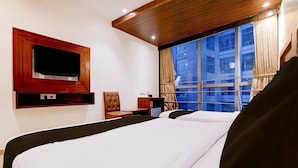 Een hotelkamer met twee bedden, een tv gemonteerd op een houten paneel, een raam met gordijnen, en een klein tafeltje met een stoel.
