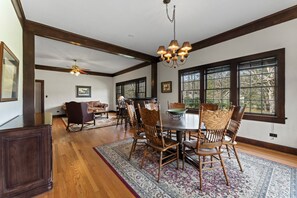 A dining area with a round table, wooden chairs, and a chandelier. There are large windows with dark frames and a view of trees outside. - Gorgeous 5-bedroom Riverside Estate Lodge in downtown Townsend (Townsend)