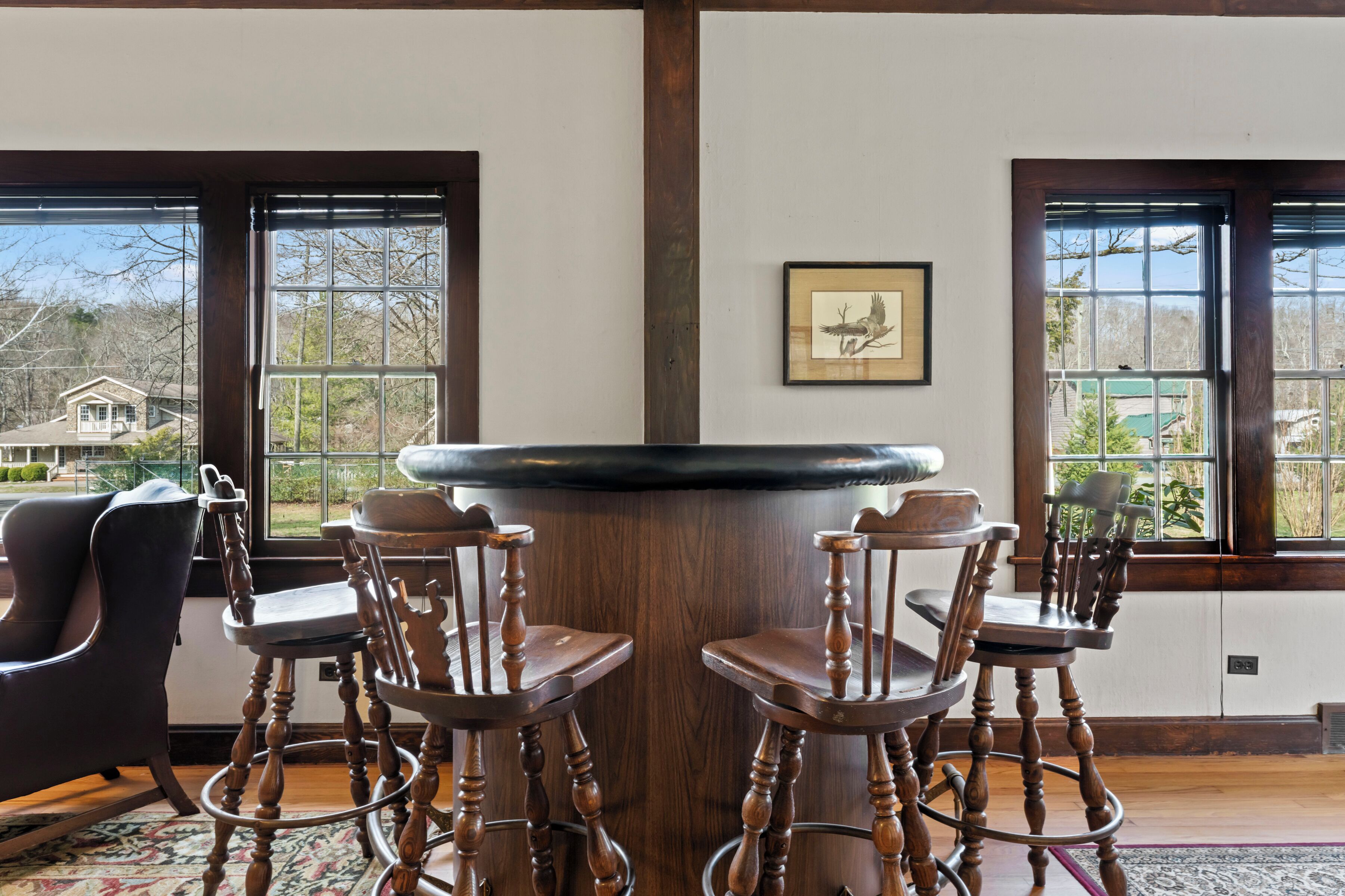 A dining area with a round table and four wooden chairs. There are two windows with views of a house and trees.