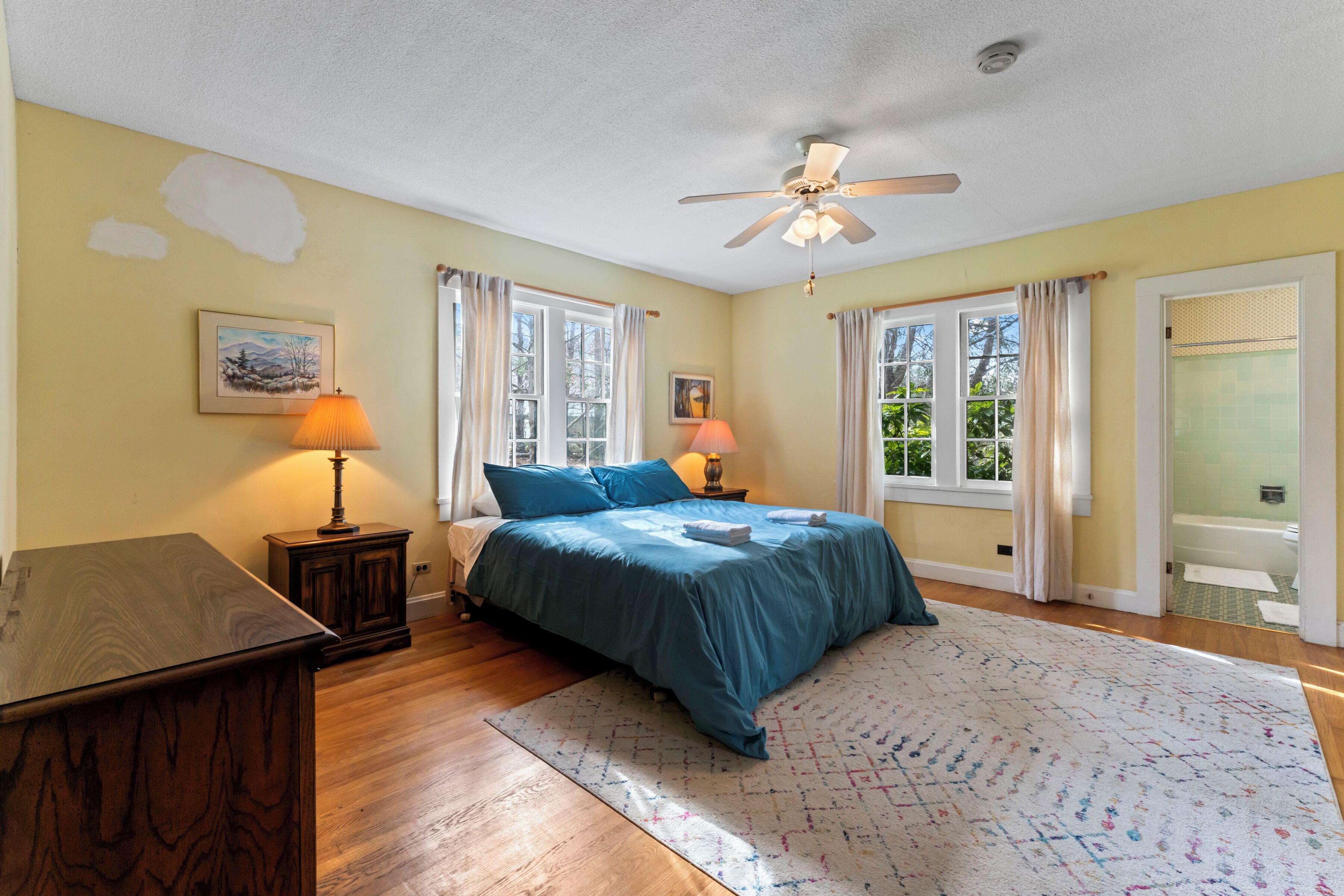 Quarto com cama, duas luminárias de cabeceira, ventilador de teto e janela com cortinas.