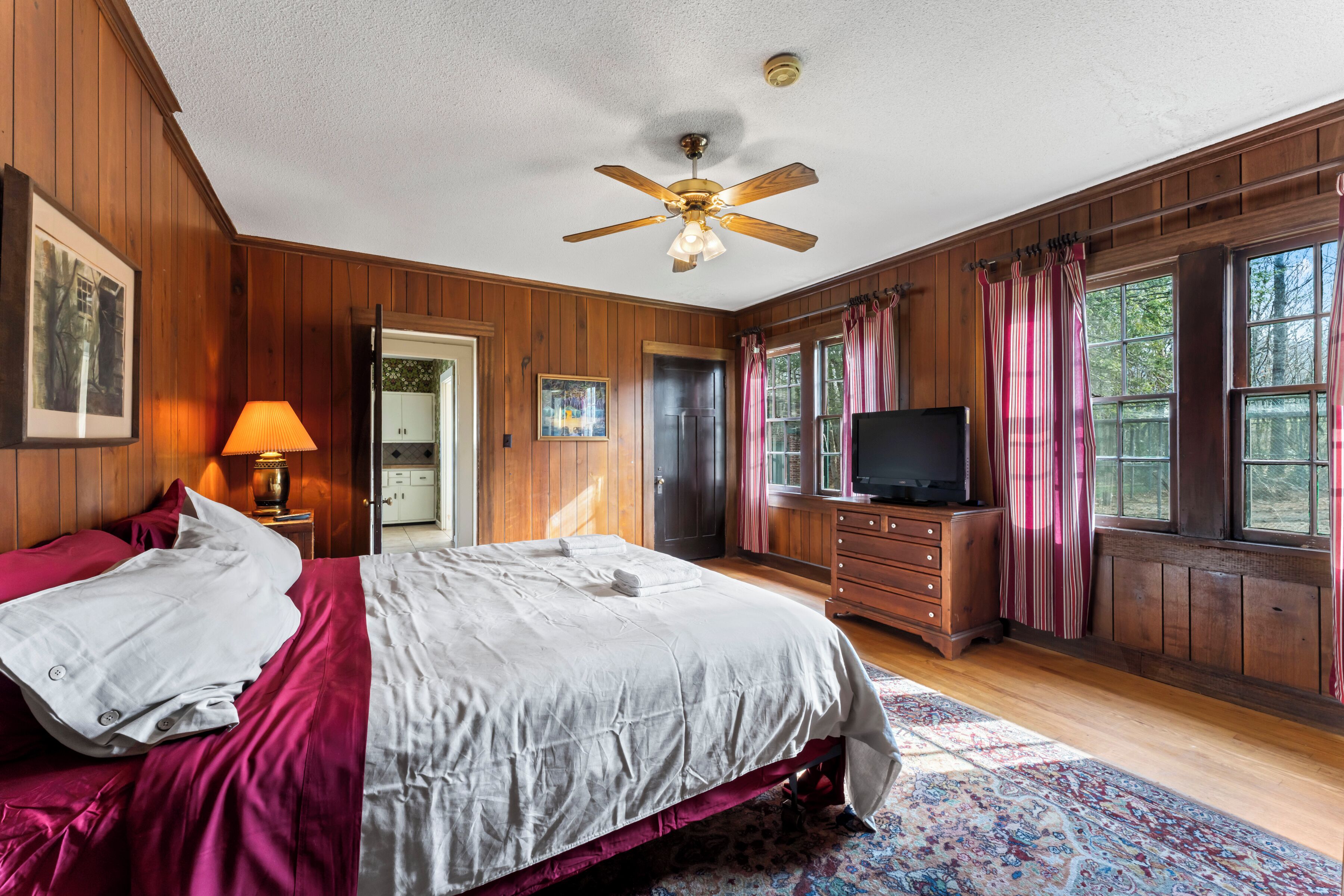 A bedroom with a bed, a television, a ceiling fan, wooden walls, and a window with curtains.