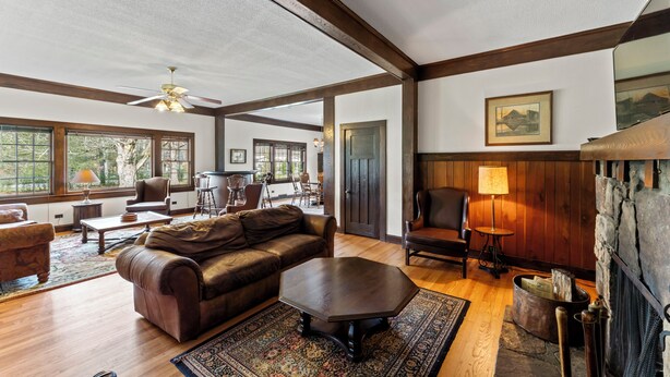 A spacious living room with a large brown leather sofa, a wooden coffee table, and a fireplace. There are multiple windows providing natural light and a view of the outdoors.